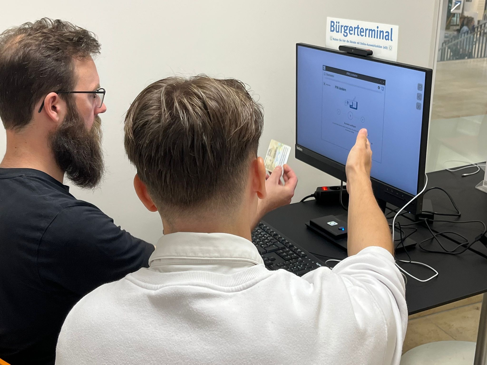 Vor einem Computer zeigt eine Person eine randere wie sie sich mit ihrem Personalausweis einloggen kann.